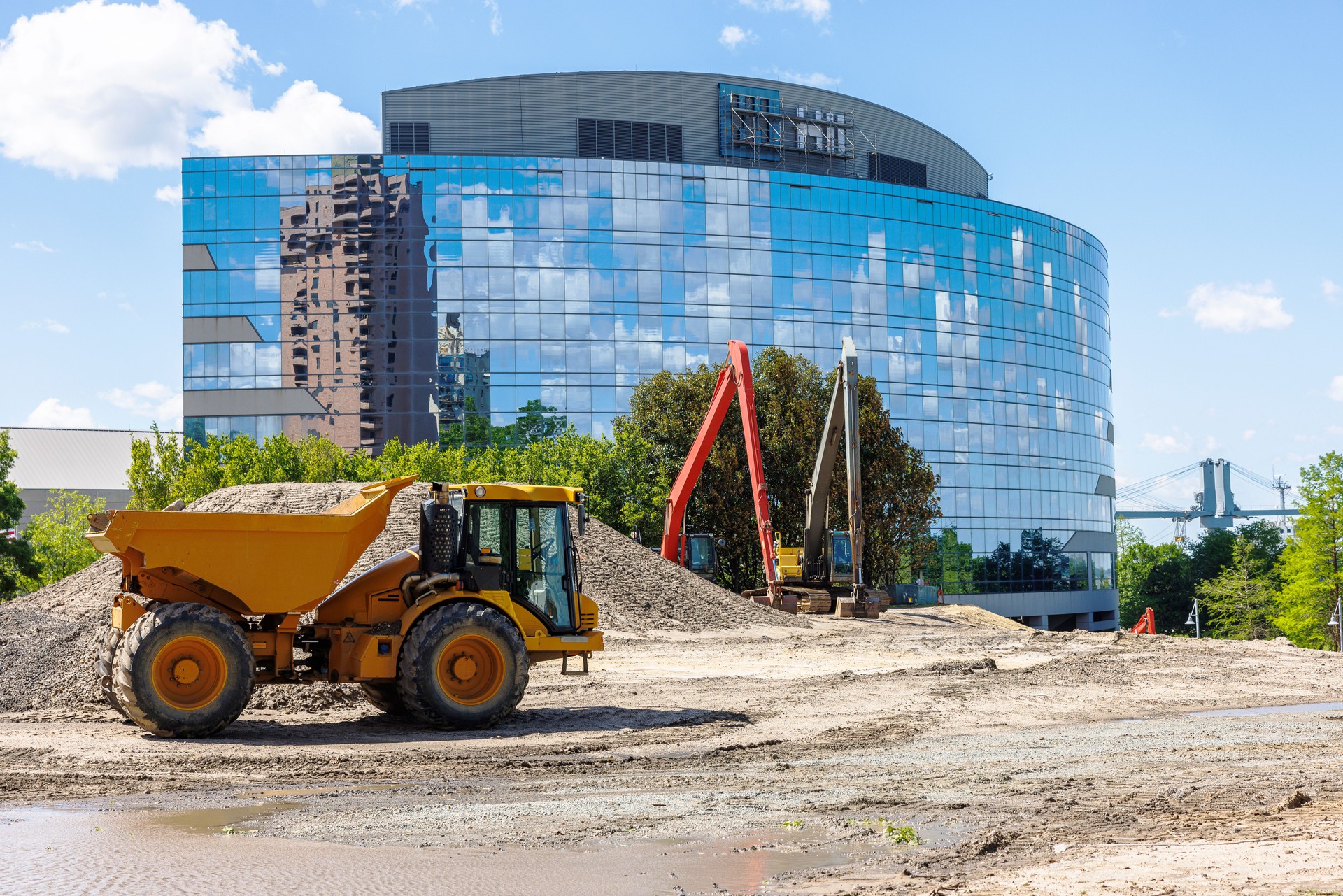 Financial district construction site in Downtown. Facade glazed office building with reflection. Dump truck and backhoe on sandy construction site against blue sky in Newport News, VA